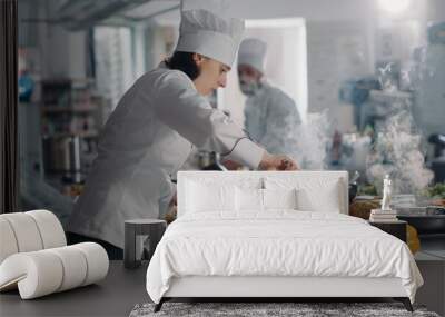 Male cook using frying pan to steam vegetables for gourmet dish, cooking delicious food with smoke in professional restaurant kitchen. Man in white uniform preparing cooked meal. Handheld shot. Wall mural