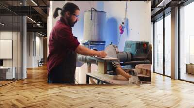 Manufacturer teaching new employee how to shape wooden bowl on disc sander in assembling shop. Apprentice learning from woodworking specialist how to use grinding machine rotating disc, camera B Wall mural