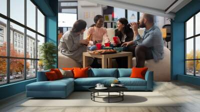 Multi ethnic colleagues having fun after work at office while eating pizza chips and drinking beer from cups and bottles. Diverse friends enjoying celebration party with snacks on table Wall mural