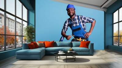 Optimistic builder looking at camera with big smile while resting after finishing work in studio shot against blue background. Contractor using tool belt needed for renovation construction tasks. Wall mural