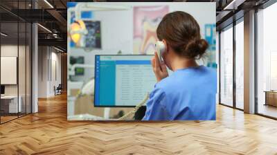 Orthodontist nurse talking at phone making dental appointments in modern equipped office, while specialist dentist with face mask is examining patient with toothache sitting on stomatological chair. Wall mural