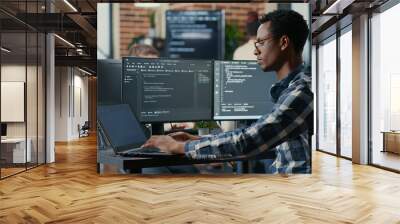 Portrait of african american developer using laptop to write code sitting at desk with multiple screens parsing algorithm in software agency. Coder working on user interface using portable computer. Wall mural