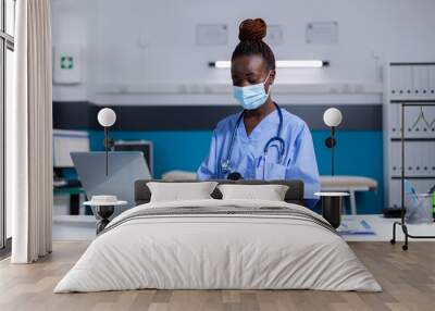 Portrait of african american nurse typing on laptop keyboard while looking at bottle of pills for medical advice. Black woman assistant wearing uniform, gloves and face mask sitting at desk Wall mural