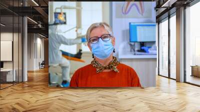 Portrait of retired patient in dental office looking on camera wearing face mask sitting on chair in waiting room clinic while doctor working. Concept of new normal dentist visit in coronavirus Wall mural