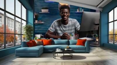 Portrait of smiling african american man using computer while working remote from home at marketing project. Young employee analyzing financial graph sitting at desk using business office technology Wall mural