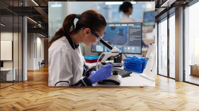 Scientist with protection glasses researching sample looking through microscope searching treatment. Medicine, biotechnology researcher in advanced pharma lab, examining virus evolution. Wall mural