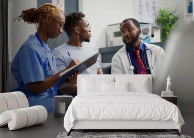 Team of clinical specialists talking to patient at hospital reception, doing medical consultation with young man. Physician and nurse explaining disease and medicare to person. Handheld shot. Wall mural