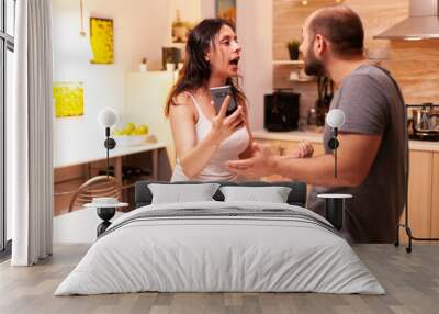 Wife angry with husband because she caught him cheating. Heated angry frustrated offended irritated accusing her man of infidelity showing him messages on smartphone yelling desperate. Wall mural