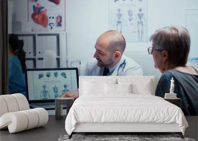 Young male doctor explaining how to avoiding bones problems to elderly retired old woman. Radiology and radiography in modern private hospital or clinic with medical staff walking in background, nurse Wall mural