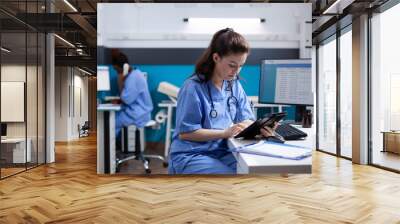 Young nurse checking appointments list on digital tablet in busy medical office. Adult woman healthcare specialist working with technology at hospital desk with stethoscope around neck Wall mural