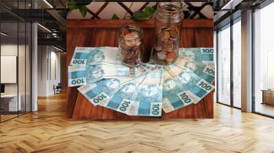 Brazilian money with several 100 reais bills and glass jars of different sizes with coins on wooden table Wall mural