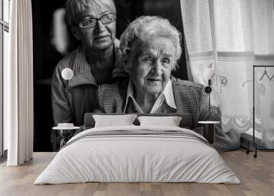 An elderly woman sitting at the table, next to a grown daughter. Black and white photo. Wall mural