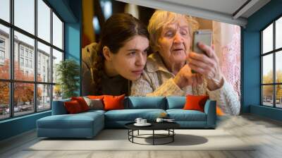 Elderly woman looks at a smartphone, with his adult granddaughter, at home. Wall mural