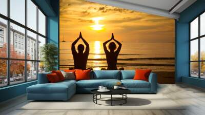 Young couple practicing yoga on the sea beach at sunset Wall mural