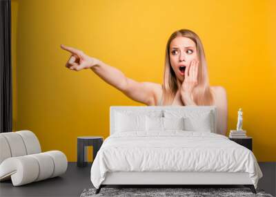 Attractive cute straight-haired blonde caucasian shocked girl, wearing casual blue shirt, pointing showing far way with finger. Isolated over yellow background Wall mural