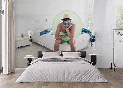 Attractive handsome young bearded sad brunet man wearing swim wear, life buoy, sunglasses, sitting indoors on bed. Summer vacation at sea resort Wall mural