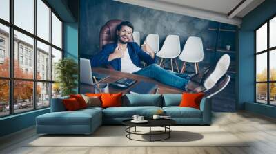 Attractive handsome young brunet bearded executive worker smiling man in office work station work place, sitting in chair, legs lying on table, dreaming, showing thumbs up Wall mural