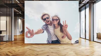 Bottom view low angle of cheerful positive joyful stylish trendy funny attractive couple in caps, casual outfits embracing showing peace, hi symbol over sky background enjoying vacation, daydream Wall mural