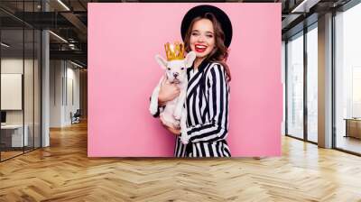 Charming, pretty, cheerful girl with beaming smile, curly hair holding dog on arms, celebrating pet's birthday, animal with gold crown on head, together looking at camera, isolated on pink background Wall mural