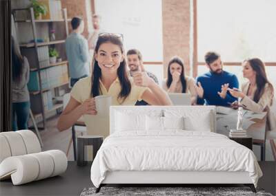 Cheerful young girl is enjoying the coffeebreak with partners, showing thumbup sign, smiling, having cup of tea, standing in casual wear in modern co working Wall mural