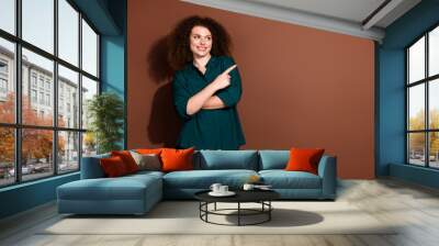 Cheerful young woman with curly brunette hair in a green shirt pointing to the side against a brown background Wall mural