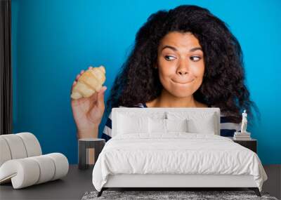 Close up photo of addicted funky afro american girl hold croissant look bite lips want eat think its yummy tasty pastry food wear stylish striped t-shirt isolated over blue color background Wall mural