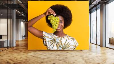 Close up portrait of cute pretty curly hair lady cover face by green grapes dress bright t-shirt isolated on yellow background Wall mural