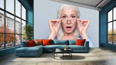 Close up portrait of stylish, aged, charming, surprised, shocked woman holding eyelets peek out glasses with wide open eyes and mouth over grey background Wall mural