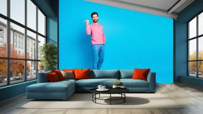 Confident young man in casual fashion with a pointing gesture, standing against a bright blue background wearing a pink sweater and jeans. Wall mural