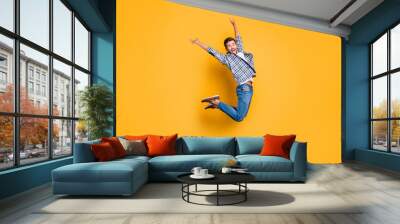 Full-body portrait of flying and cheerful man in sneakers, denim outfit, jumping with raised arms isolated on shine yellow background Wall mural