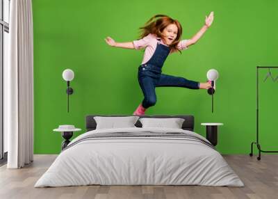 Full length photo of cheerful funny pretty little schoolchild jumping high fighting exercises training wear casual denim overall pink shirt isolated green background Wall mural