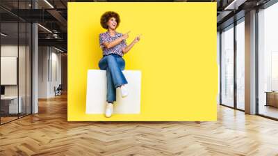 Full length photo of cute dreamy woman wear print shirt sitting cube showing two fingers empty space isolated yellow color background Wall mural