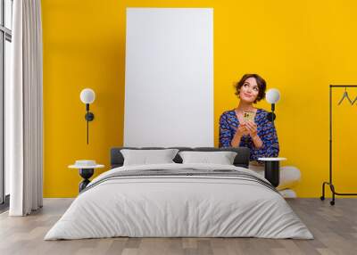 Full length photo of dreamy cute lady looking at phone touchscreen with device promoting shopping app isolated on yellow color background Wall mural