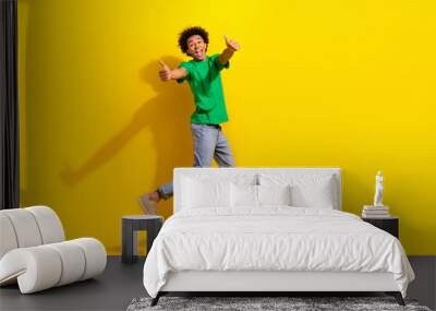 Full length photo of funky cool guy dressed green t-shirt jumping high showing two thumbs up emtpy space isolated yellow color background Wall mural