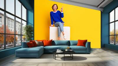 Full length photo of funky dreamy girl wear blue pullover sitting podium pointing empty space isolated yellow color background Wall mural