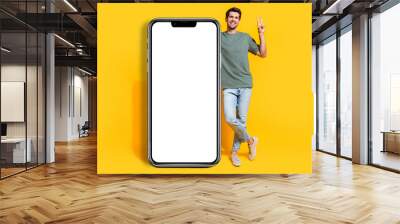 Full length photo of good mood cute guy wear khaki t-shirt showing v-sign modern device empty space isolated yellow color background Wall mural