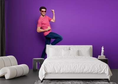 Full length photo of handsome attractive guy beaming smiling rejoicing party started raise fists wear sun glasses casual pink t-shirt jeans isolated purple color background Wall mural