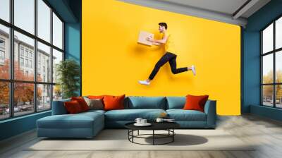 Full length photo of handsome guy jumping high holding big parcel box hurry move new apartments wear casual t-shirt pants isolated yellow color background Wall mural