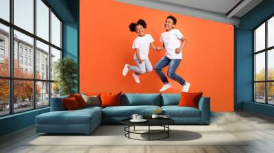 Full length photo of little funky girl boy jump wear white t-shirt jeans sneakers isolated on orange background Wall mural