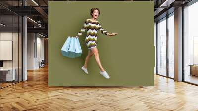 Full length photo of pretty cute girl dressed striped clothes jumping high holding shoppers empty space isolated green color background Wall mural