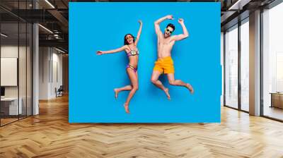 Full length portrait of a joyful young jumpers woman and happy man in sunglasses, dressed in swimsuit, jumping and putting hands up over blue background. Beach party style! Wall mural