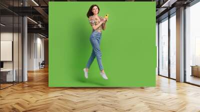 Full length portrait of charming energetic person jumping use smart phone empty space isolated on green color background Wall mural