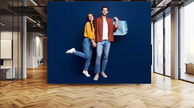 Full length portrait of cheerful funny partners hold hands carry store bags isolated on blue color background Wall mural