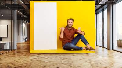 Full length portrait of cheerful person sit floor hold telephone direct thumb finger empty space board isolated on yellow color background Wall mural