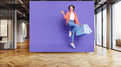 Full size photo of nice young girl hold bags jump direct finger empty space wear shirt isolated on violet color background Wall mural