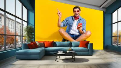 Full size photo of pleasant guy wear jeans jacket sit hold smartphone indicating at offer empty space isolated on yellow color background Wall mural