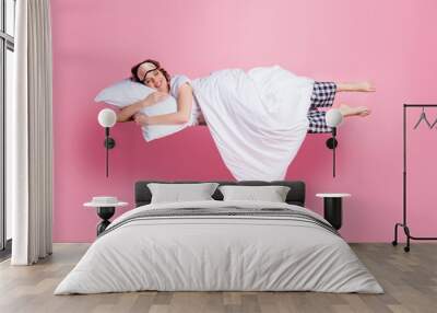 Full size photo of pretty cheerful lady satisfied morning nap lying bed hug pillow covered blanket wear mask white t-shirt plaid pajama pants barefoot isolated pink color background Wall mural