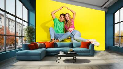 Full size photo of two nice young people sitr floor arms make roof gesture isolated on bright yellow color background Wall mural