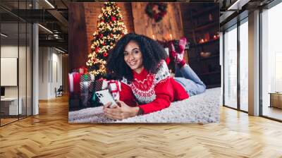 Full size portrait of positive nice girl lying carpet floor hols smart phone texting cozy christmas atmosphere tree garland indoors Wall mural