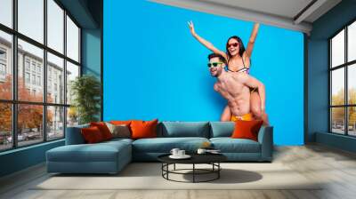 Handsome man wear in summer glasses and yellow shorts keep on back charming young woman holding hands to side making airplane isolated on blue background Wall mural